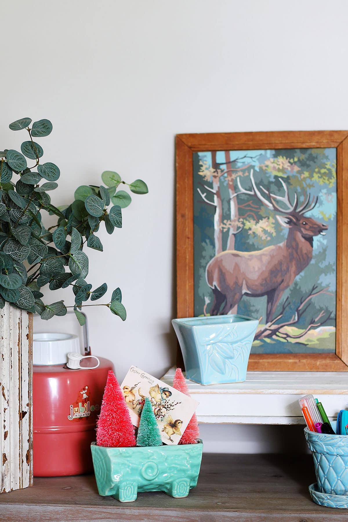 A printable Easter image tucked in a planter being used as spring decor. A colorful vintage paint by number painting of a deer is in the photo also.