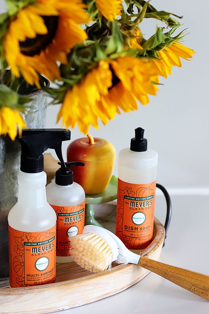 Mrs. Meyer's fall scents including Mrs Meyer's hand soap, dish soap and multi-surface spray setting on a wooden tray with yellow sunflowers in a bucket.