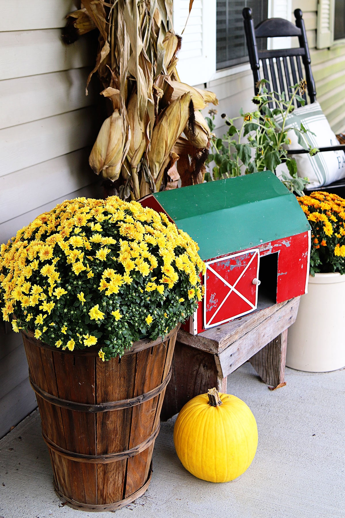 Fall porch decor with a yellow pumpkin, bushel basket with a yellow mum and a handmade toy sized red barn.