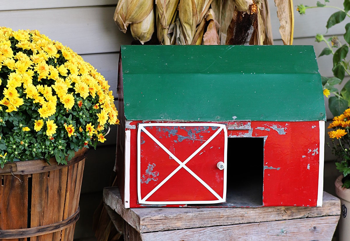 A handmade folk art toy barn setting on a porch decorated in fall decor.