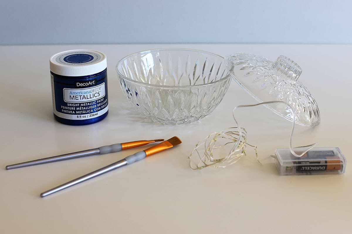 supplies for painting glassware laid out on the table - paint, brushes, battery operated lights and a pressed glass candy dish.