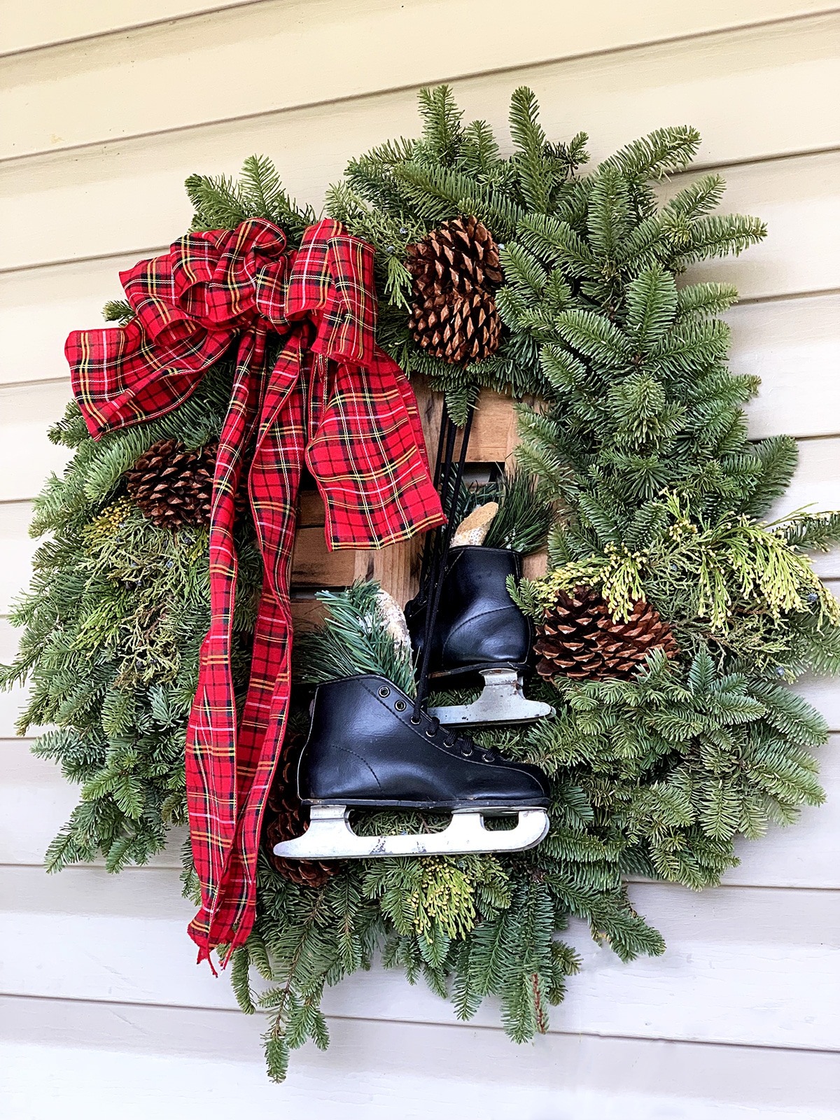 A winter wreath for a Christmas front porch - with pine boughs, pinecones, tartan plaid bow and black ice skates.