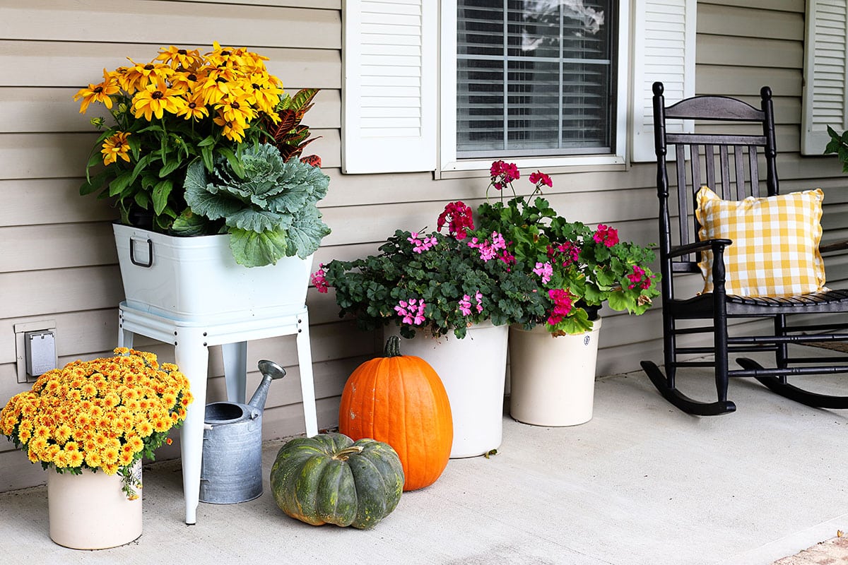 A simple fall porch with fall flowers in a wash tub and black porch rockers with yellow pillows. 