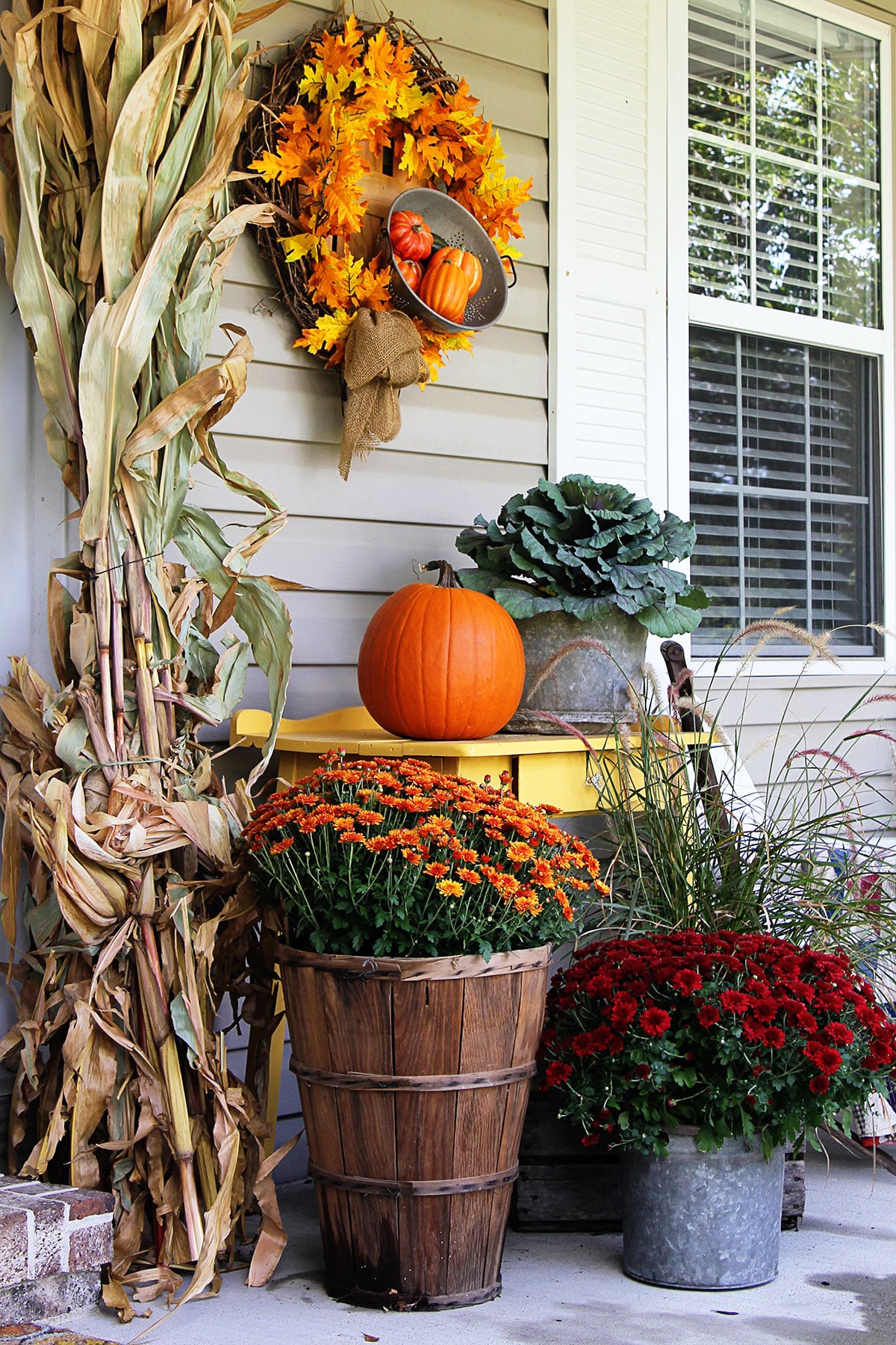Fall porch decorations including orange  and red mums, ornamental cabbage, corn stalks and an orange pumpkin.