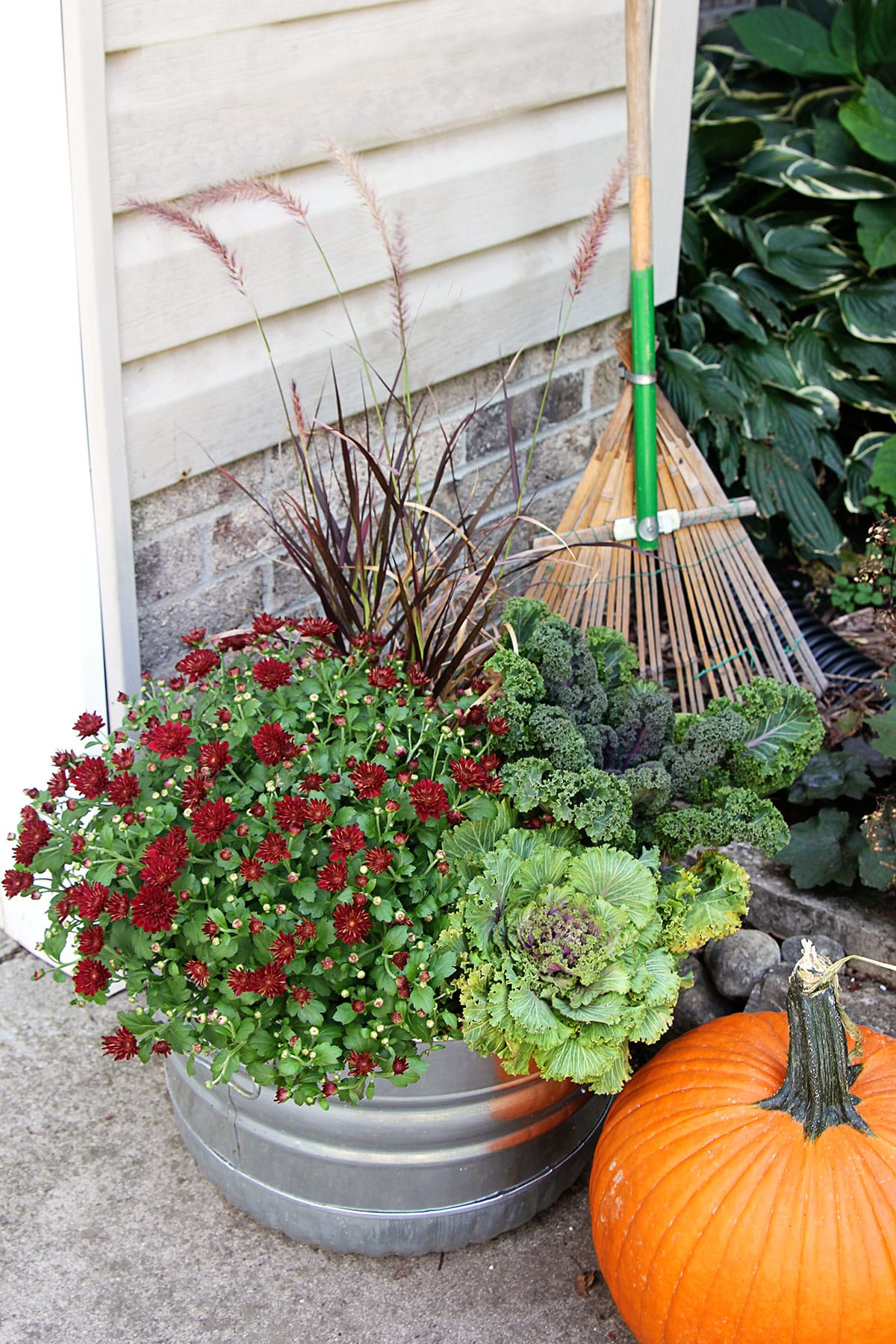 Fall plants in a galvanized tub with a vintage garden rake and an orange pumpkin.