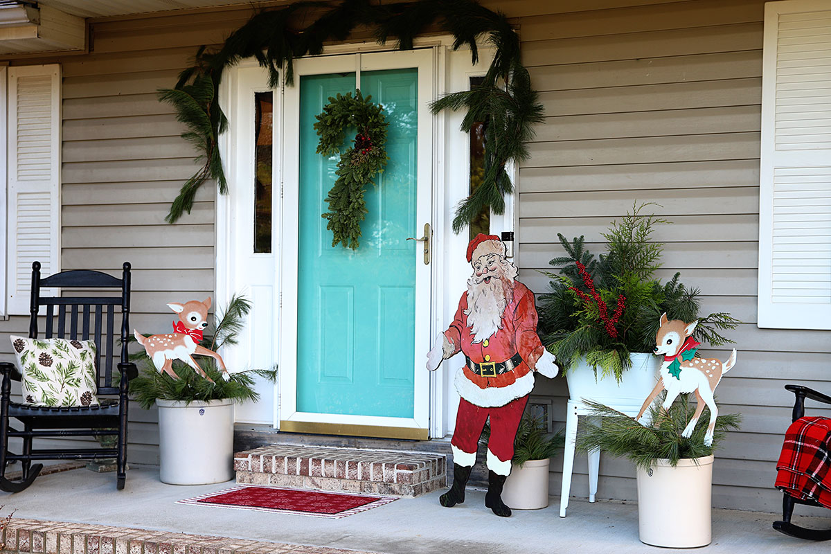 Front porch decorated for Christmas with lots of evergreen garland, wreath and vintage inspired characters - reindeer and Santa.