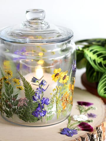 Glass jar decoupaged with dried pressed flowers setting on a wooden tree slab.