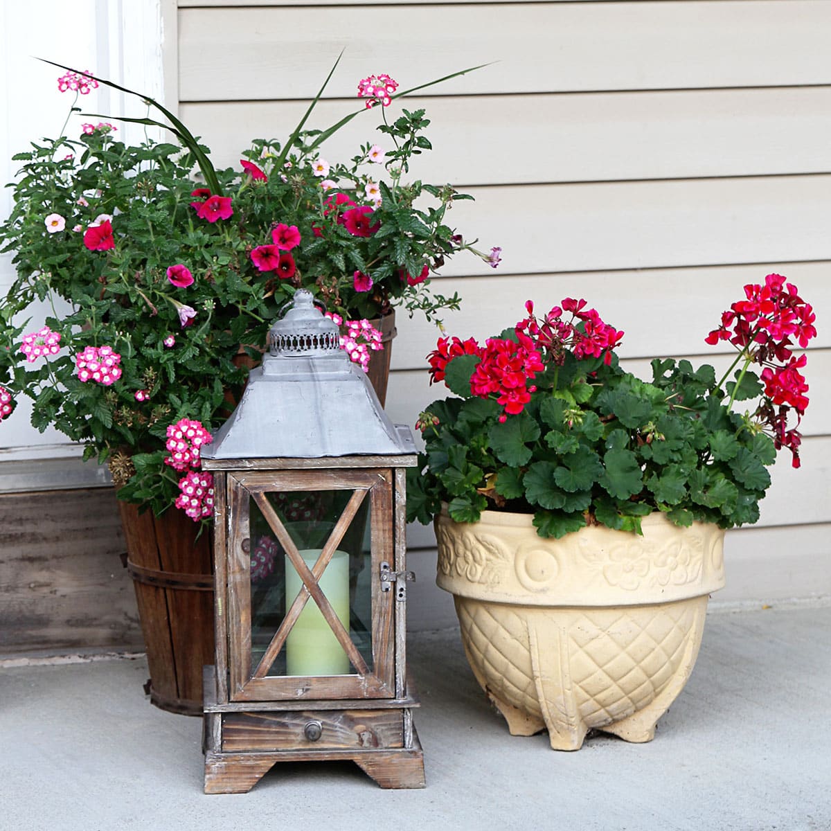 Vintage stoneware planter with red geraniums planted in it. Setting next to a bushel basket of flowers and a wooden farmhouse lantern.