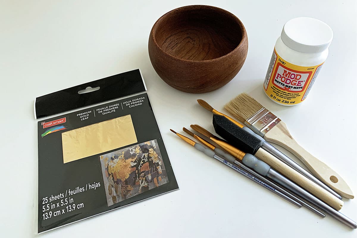 Gold leaf sheets, various craft paint brushes, Mod Podge and a wooden bowl all of which will be used to gold leaf the wooden bowl.